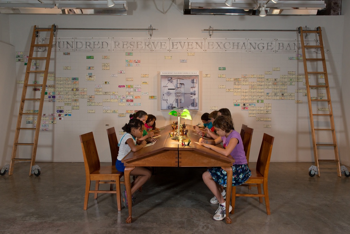 Students draw Fundreds on the handcrafted cherry tables in the Charlotte branch of the Fundred Reserve Even Exchange Bank, 2013. [Fundred Project, 2008-2019.] Photo: Ben Premeaux, courtesy Fundred Project.